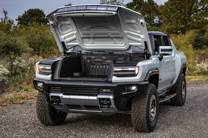 Electric SUV with hood raised, showcasing the engine compartment, parked on a gravel road surrounded by trees.