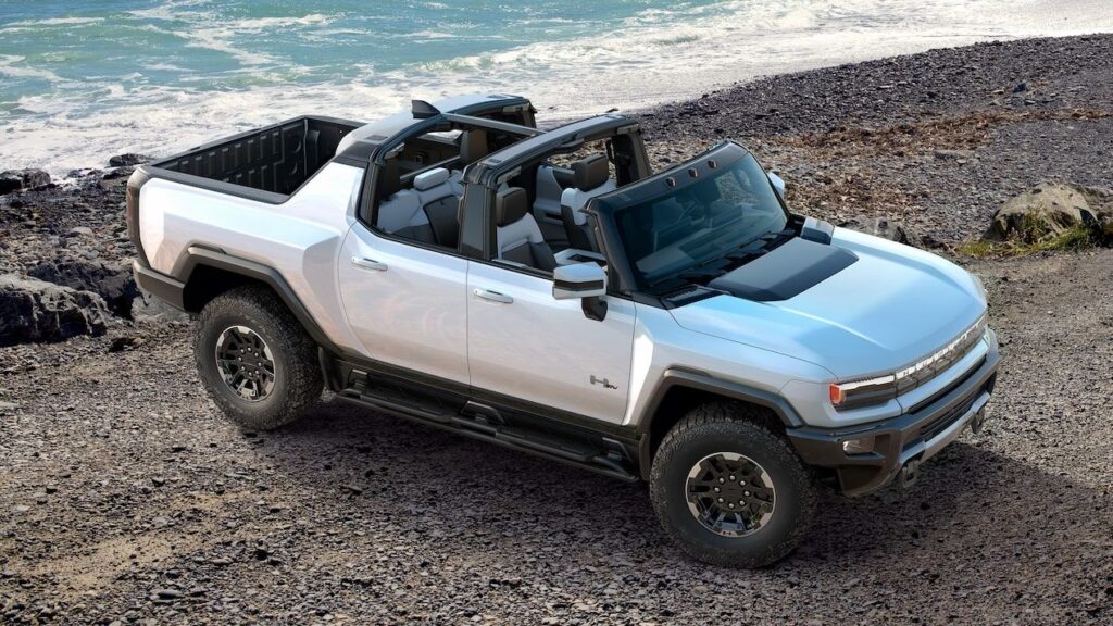All-terrain electric pickup truck parked on rocky shore near ocean, showcasing a sleek silver exterior, open roof, and spacious interior with modern design elements.