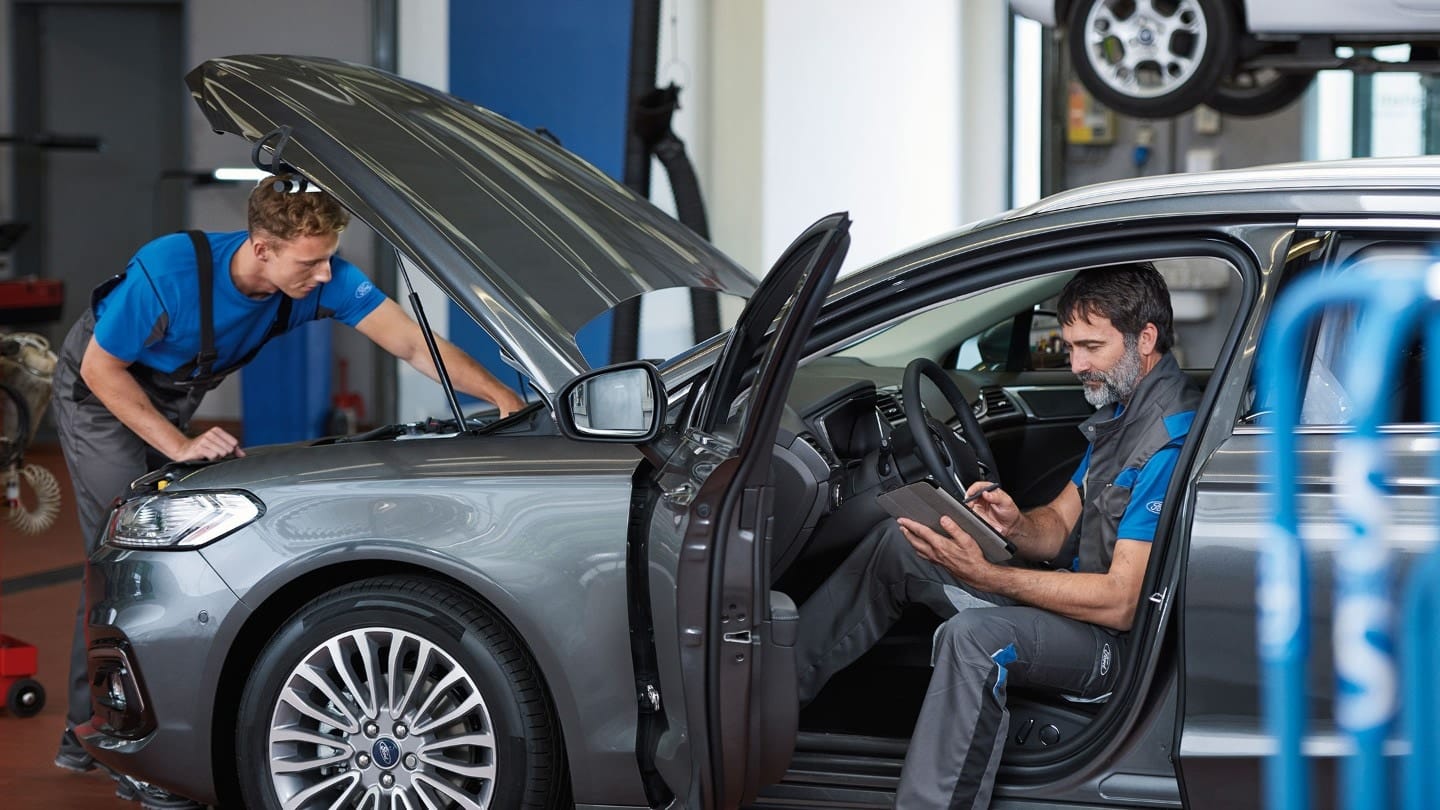 Two mechanics working in a modern auto repair shop; one examining the engine of a grey car with its hood open, while the other sits inside the vehicle, using a tablet to check diagnostics.