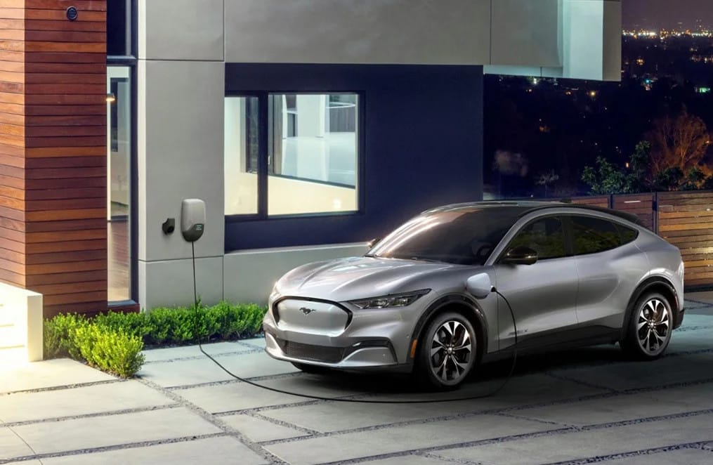 A sleek silver electric vehicle parked in a modern home garage, charging from a wall-mounted electric car charger, surrounded by landscaped greenery at dusk.