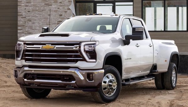 New Chevrolet Silverado HD 2023 model in white color showcasing a robust front grille and streamlined design, parked on sandy terrain with a modern building in the background. Ideal for heavy-duty performance and outdoor adventures.
