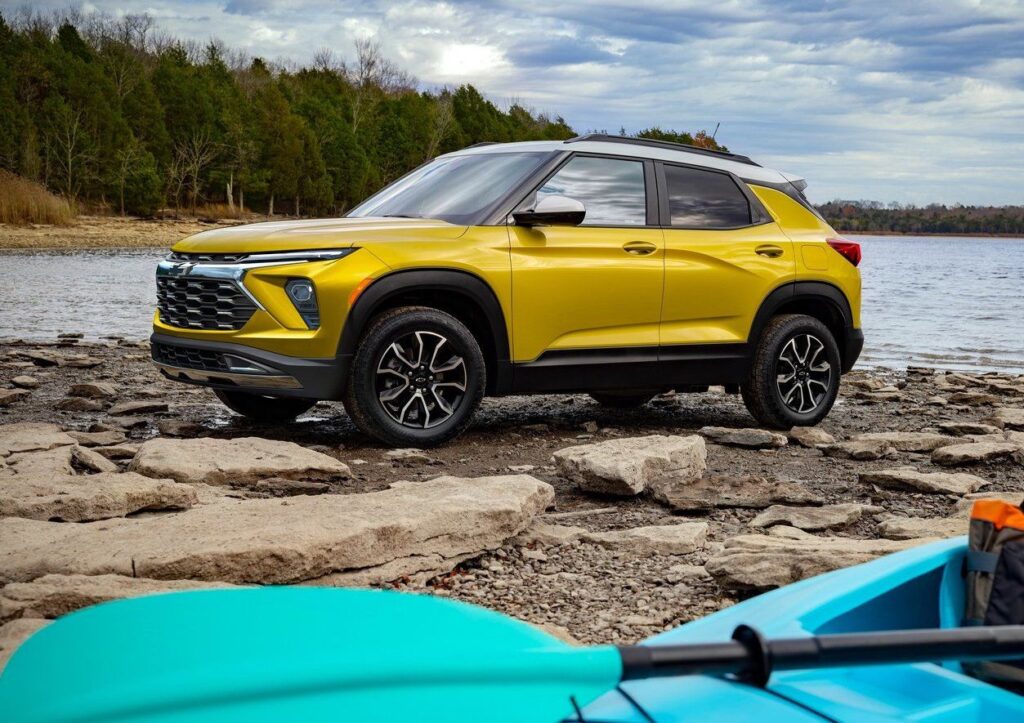 Yellow compact SUV parked on rocky shoreline near a lake, with trees in the background and a kayak visible in the foreground. Ideal for outdoor adventures, showcasing rugged design and versatility.