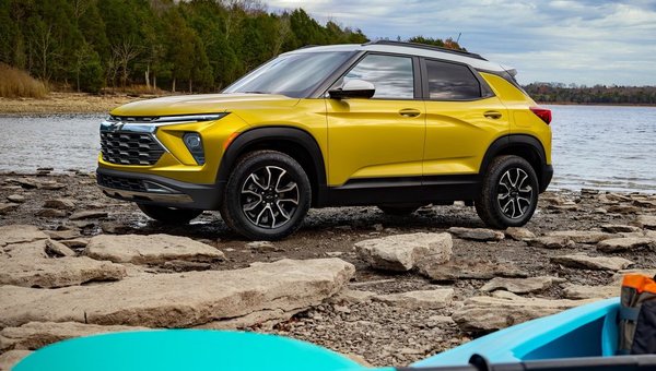 A vibrant yellow SUV parked on rocky terrain near a lake, surrounded by trees, showcasing its modern design and features under a cloudy sky.