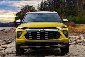 Front view of a bright yellow Chevrolet SUV parked on rocky terrain near a lake, showcasing its modern design and sleek lines against a backdrop of trees and a cloudy sky.