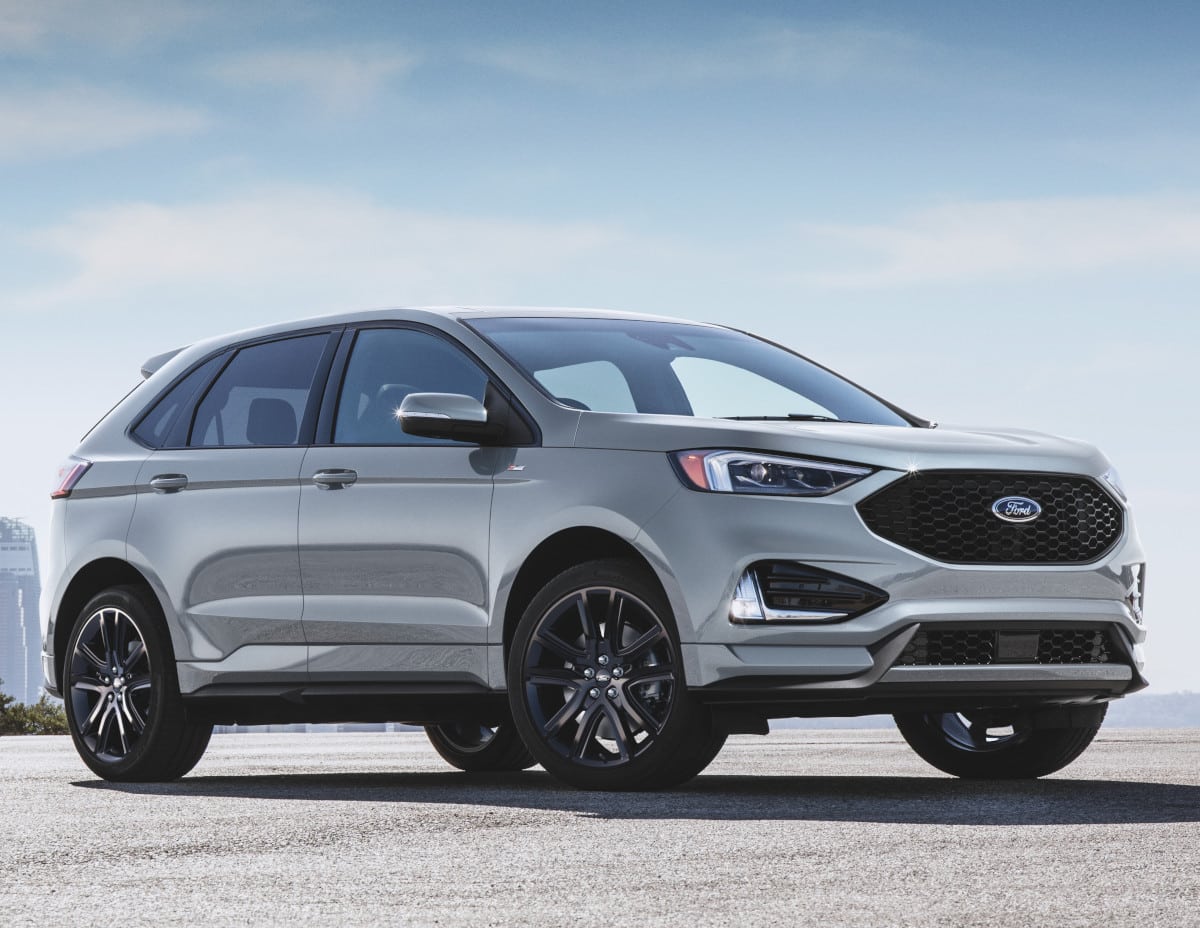 Ford Edge SUV in silver with a modern design and sporty black wheels, parked against a clear blue sky background. Ideal for families and adventure seekers, showcasing its sleek profile and high-quality finish.