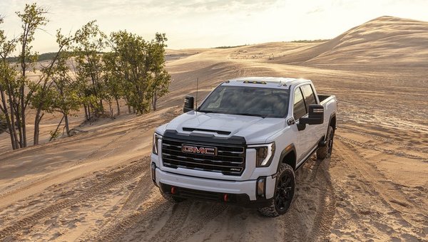 White GMC truck parked on sandy dunes under a clear sky, showcasing rugged design and off-road capabilities surrounded by trees.