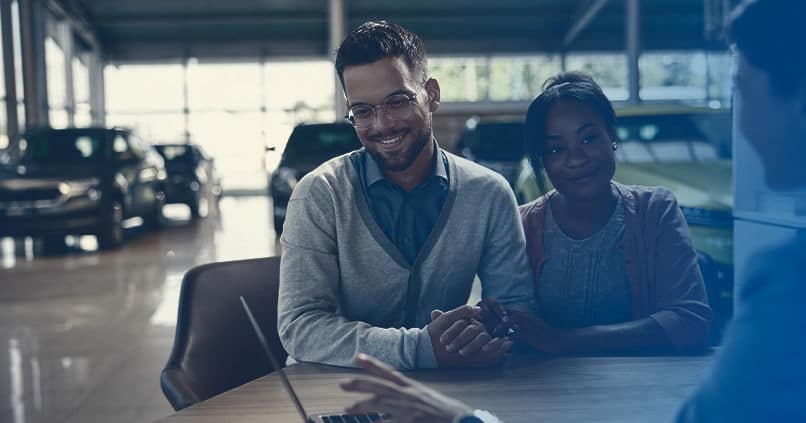 A smiling couple engages in a discussion with a car salesman at a dealership, exploring options for purchasing a vehicle. The background shows various cars on display, creating an inviting atmosphere.