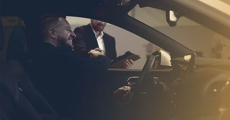 A smiling couple engages in a discussion with a car salesman at a dealership, exploring options for purchasing a vehicle. The background shows various cars on display, creating an inviting atmosphere.
