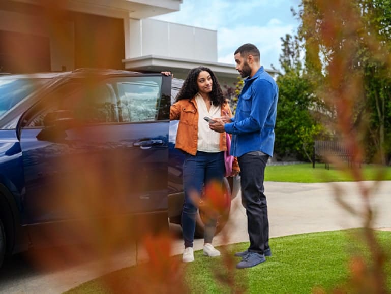 A couple discussing car details while standing beside a blue SUV parked in a driveway, surrounded by greenery and a modern home.