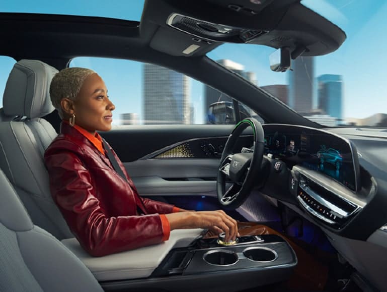 Interior view of a luxury car featuring a woman with short hair in a stylish outfit, seated in the driver's seat. The dashboard displays advanced technology and a panoramic city skyline can be seen through the windshield.