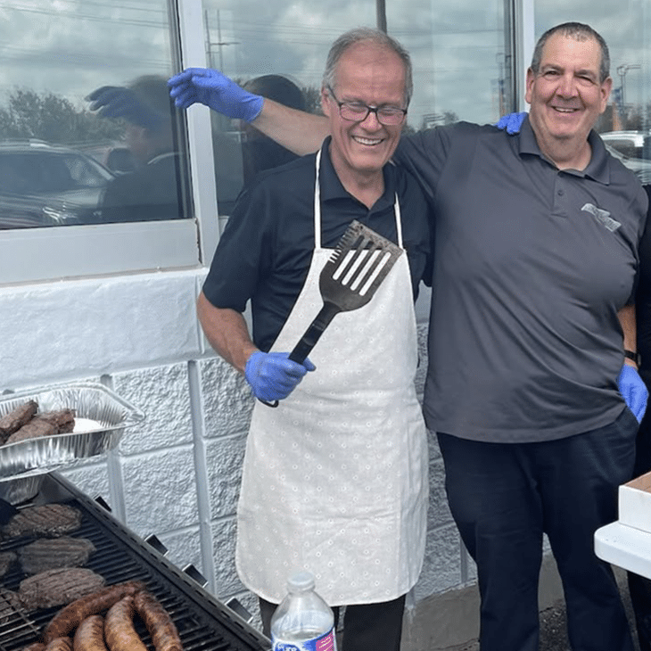 Two smiling men at a barbecue, one holding a spatula, with grilled food in the foreground.